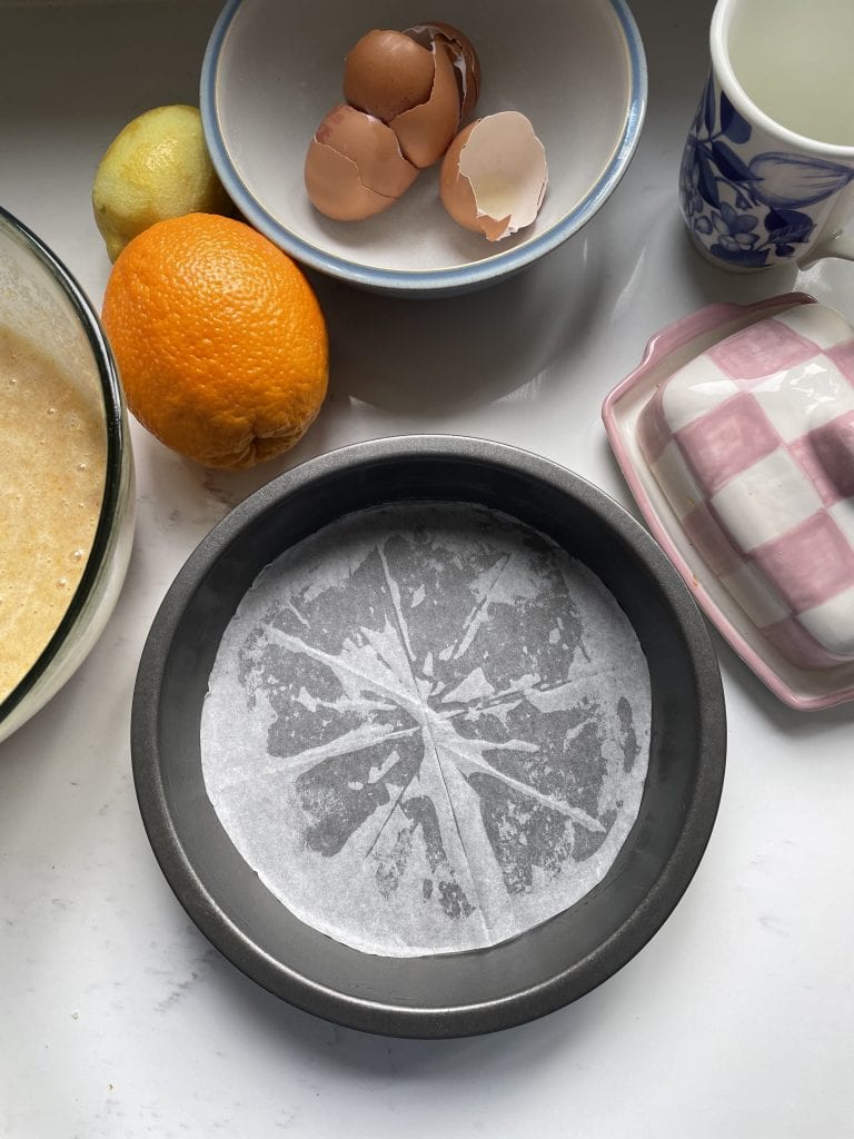 Lined cake tin surrounded by ingredients for baking an orange cake.